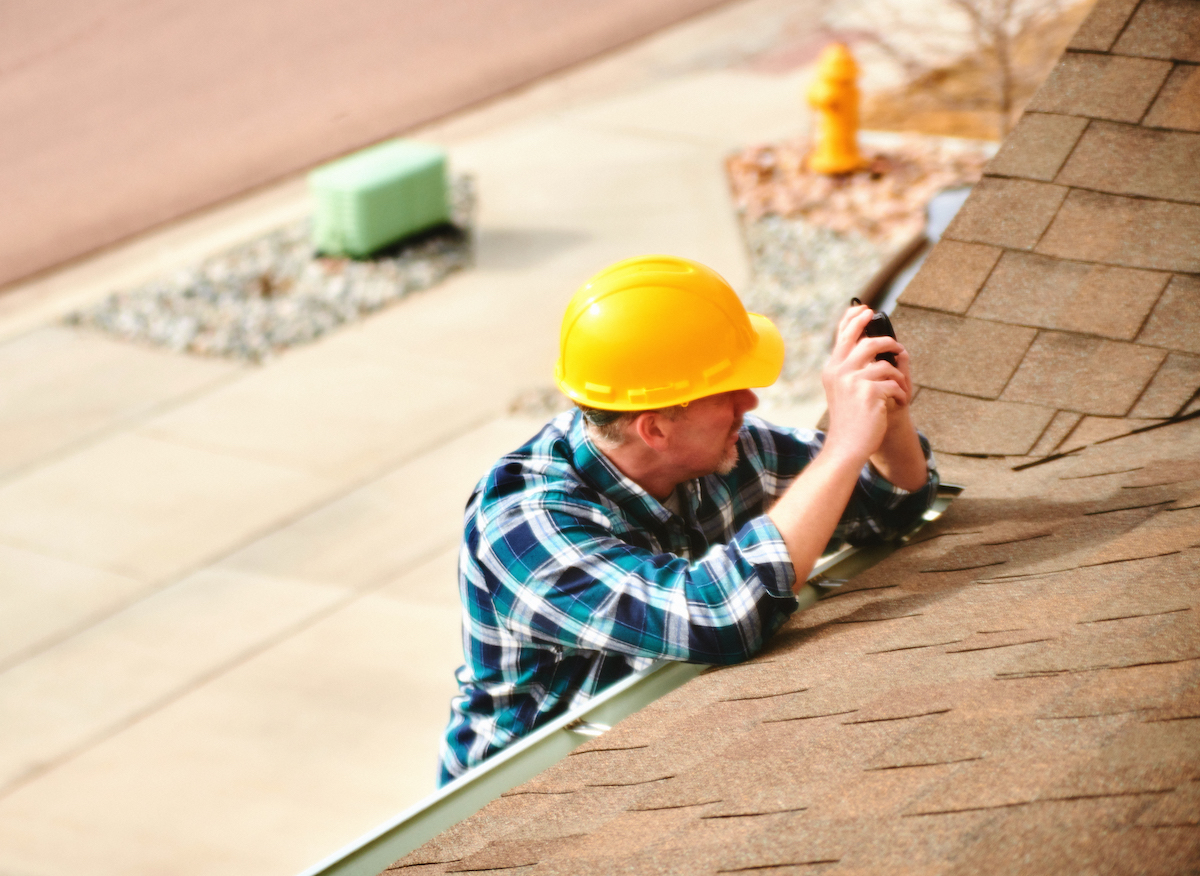 Roof appraiser or agent with hard hat peers over side of house to roof to examines status of brown roof.