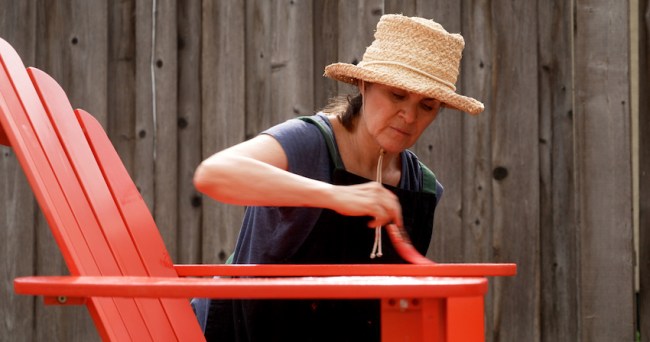Woman wearing hat outdoors painting an Adirondack chair red.
