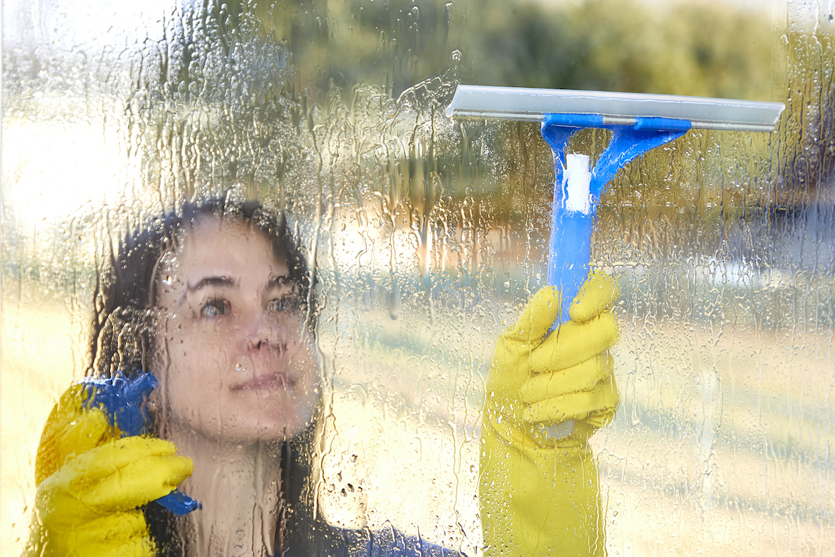 Woman wearing yellow rubber gloves washing exterior windows with a spray and squeegee.