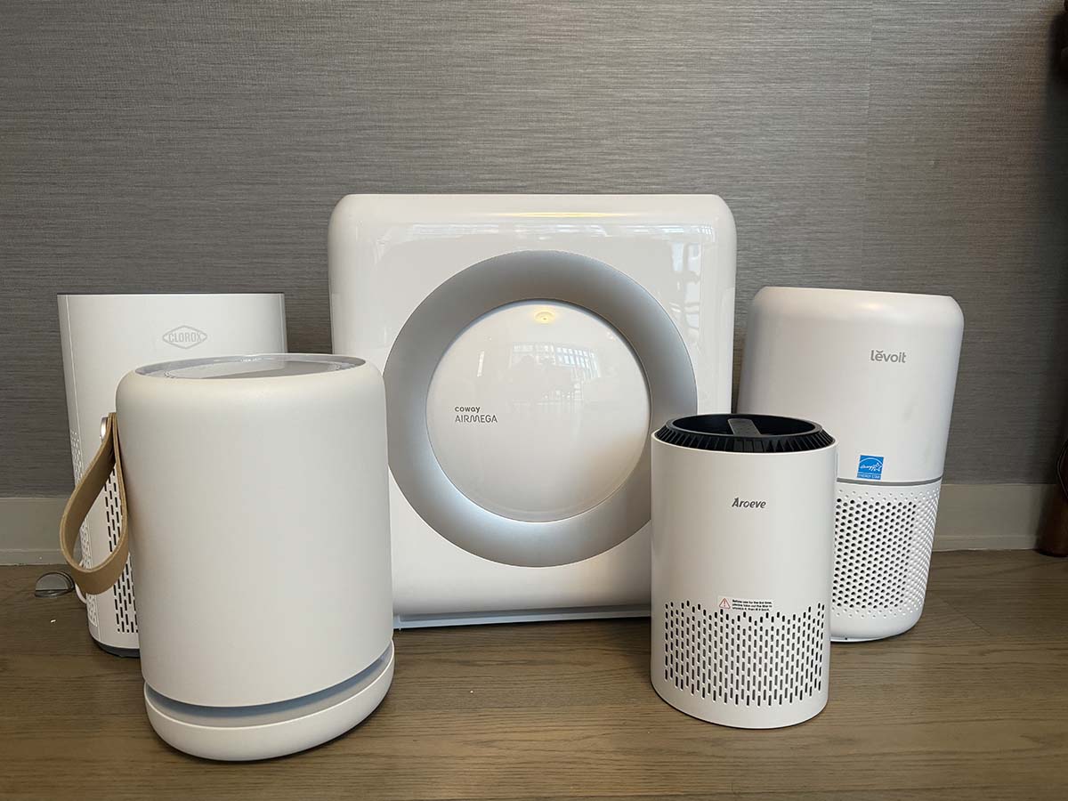 Five white and gray air purifiers on a wood floor in front of a gray wall