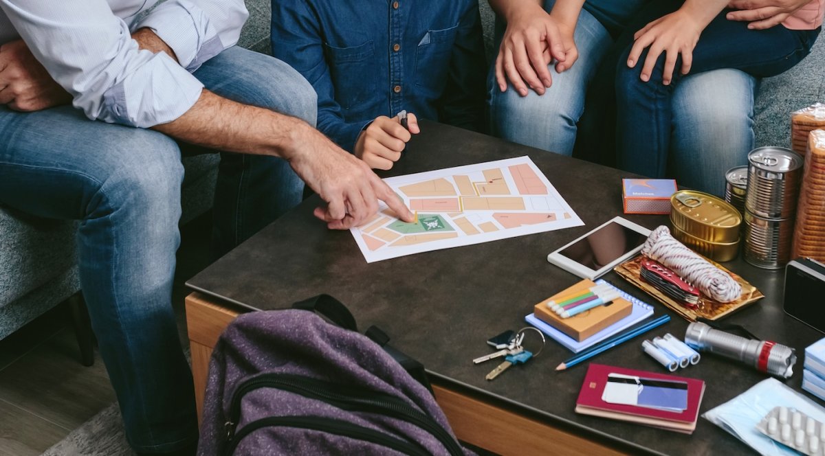 Father showing family an emergency evacuation map as emergency supplies and food stand on the coffee table in front of them.