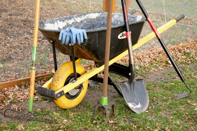 Rake, shovel, gloves, and other gardening tools assembled on a wheelbarrow outdoors.