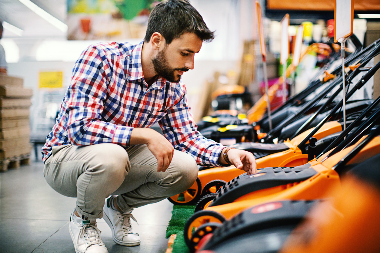 mowing mistakes everyone makes bearded man kneeling to look at lawn mower shopping hardware store