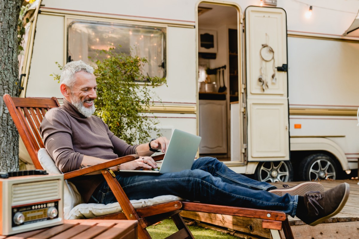 iStock-1282881814 camper decor Attractive grey-haired man resting on the wooden deck chair using laptop with caravan van behind