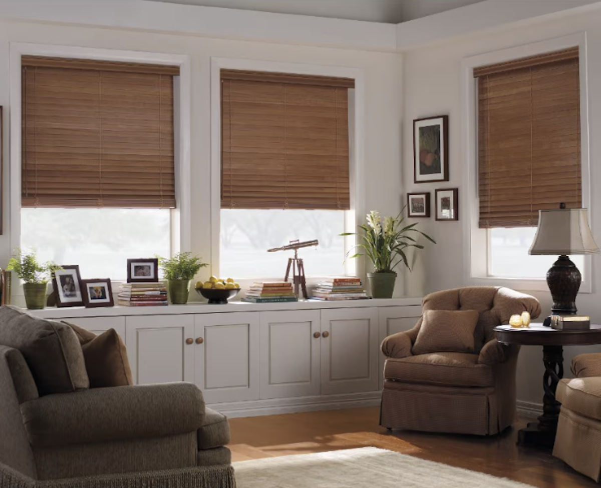 A bright living room with white built-ins and tan armchairs with large windows dressed in wood blinds. 