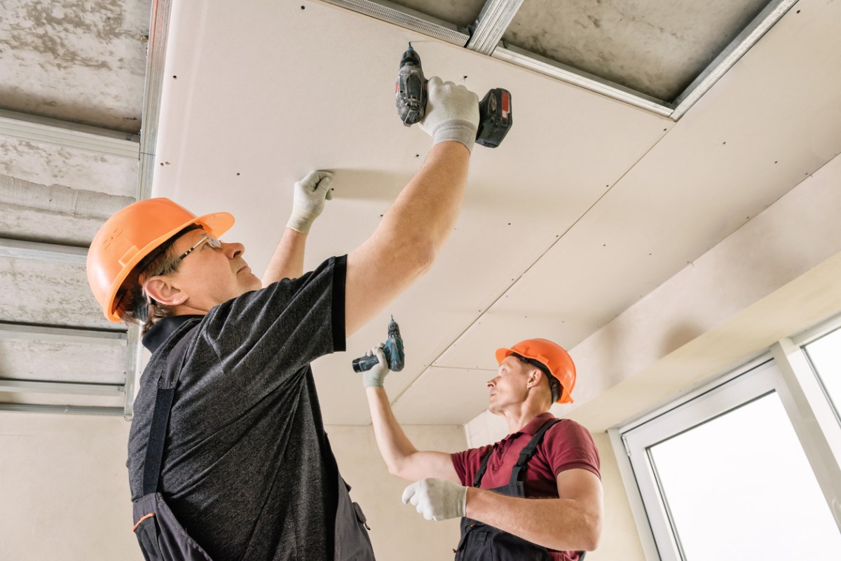 Two men wearing hard hats screw drywall into a ceiling.