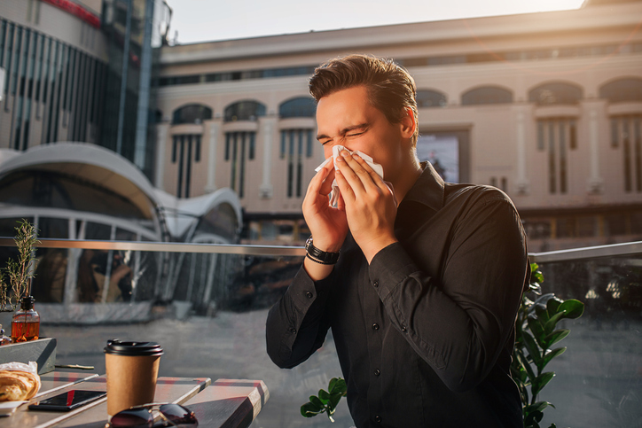 young-man-in-black-dress-shirt-sneezes-into-a-tissue-while-sitting-in-a-patio-among-buildings-in-a-city