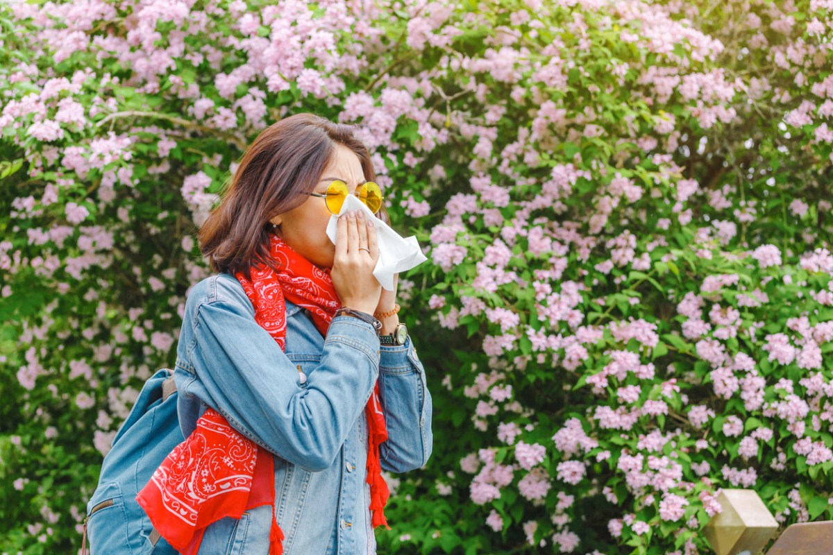 young woman with backpack and red scarf sneezes with tissue in front of large flowering tree