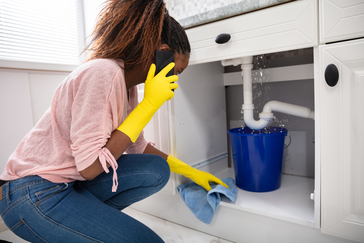 a-woman-in-pink-shirt-and-jeans-wears-yellow-rubber-gloves-while-making-a-phone-call-and-looking-at-a-leak-under-the-sink-caught-by-a-bucket