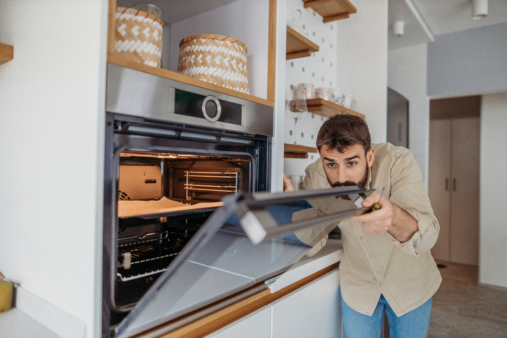 a-bearded-man-in-a-tan-shirt-holds-a-screwdriver-while-he-looks-inside-a-wall-mounted-stove-in-a-white-kitchen