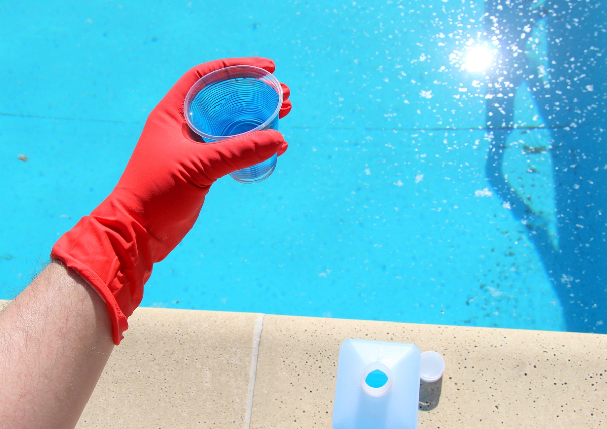 uses for hydrogen peroxide - red gloved hand holding cup of solution over swimming pool