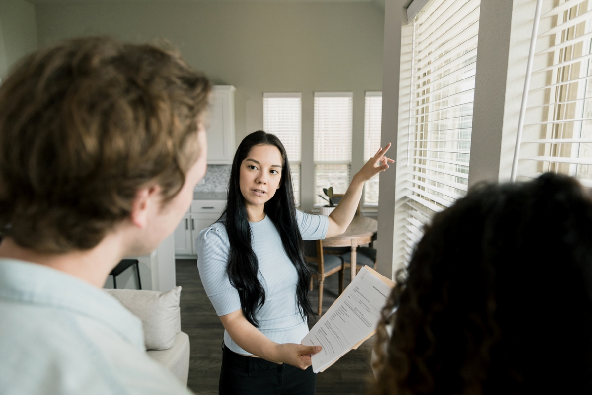 Young woman holding checklist and explaining home features to blurred couple