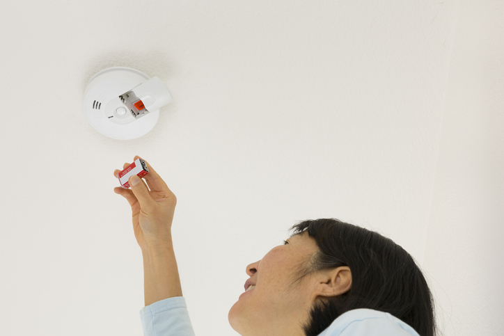 a-woman-reaches-up-to-change-the-battery-in-a-smoke-detector-on-a-white-ceiling