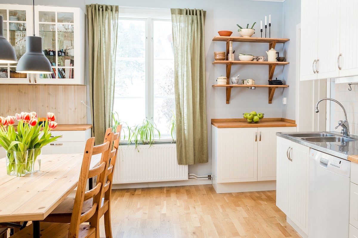 Sunllit kitchen with a wooden table and green apron-length curtains on the window.