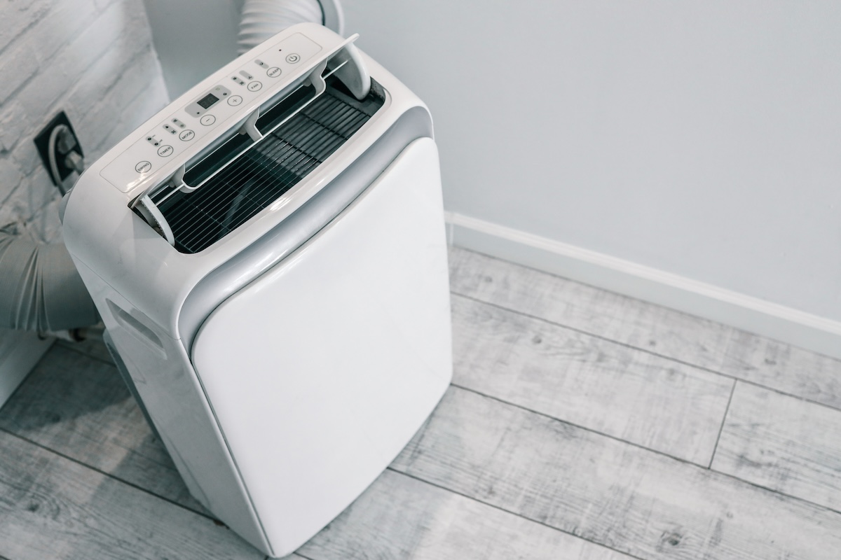 A close up of portable air conditioner with digital display and control panels placed on a wooden floor near the wall socket.