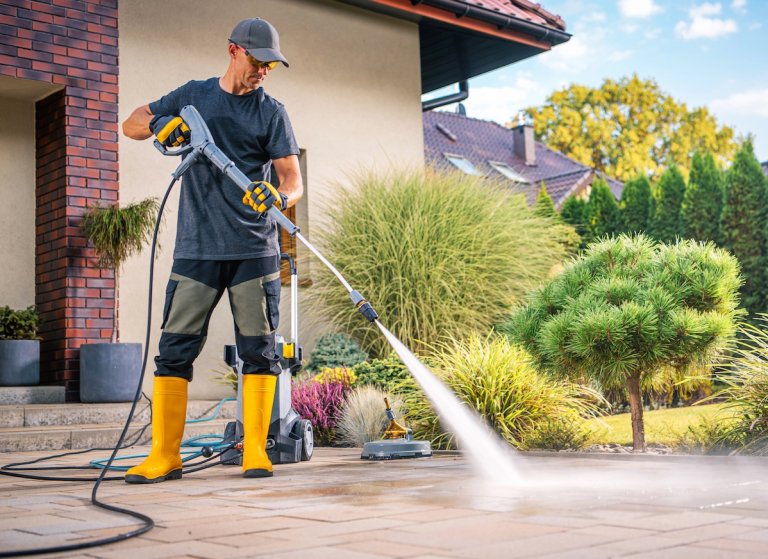 A young man pressure washes the patio in front of a nicely landscaped stucco house.