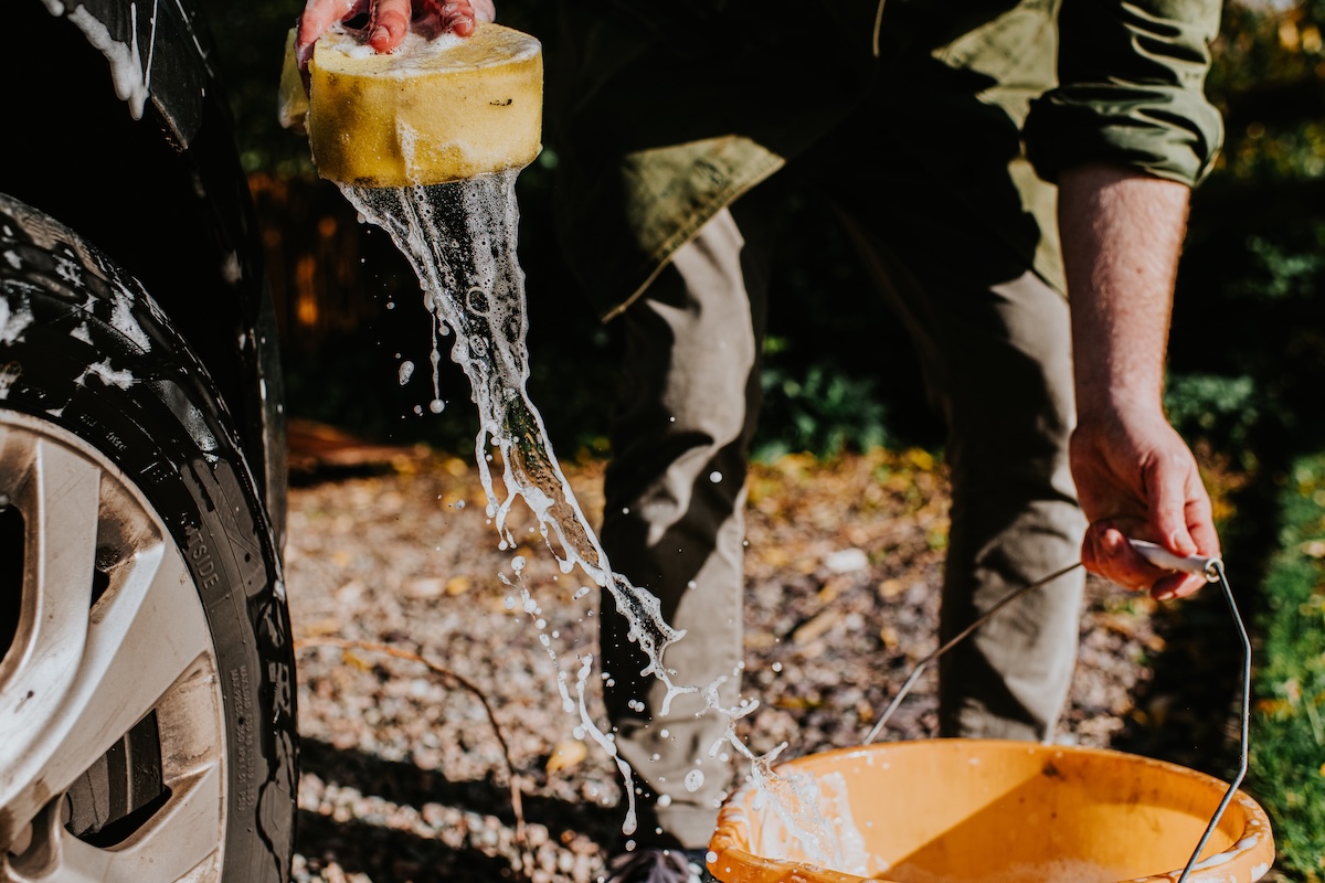 A man bends down to wash a car, using a sponge and bucket filled with soapy water.
