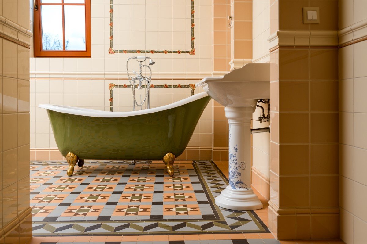 A vintage-styled bathroom featuring a green and white clawfoot freestanding bathtub paired with a decorative pedestal sink and patterned geometric tile flooring.