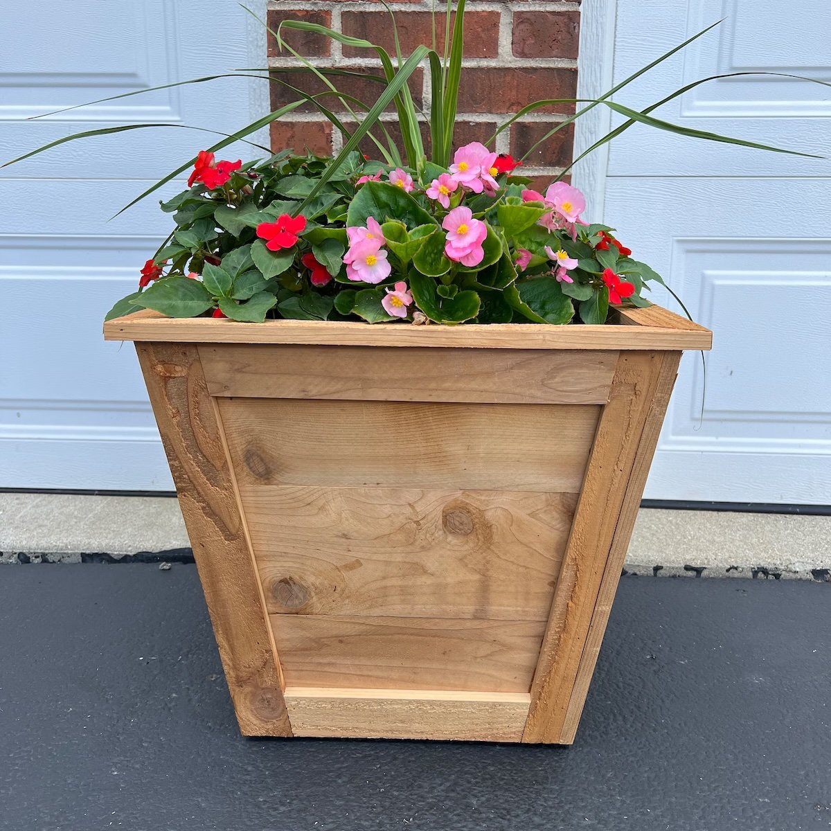 Cubical wooden planter is full of red and pink flowers and sits in front of a brick wall and white garage door.