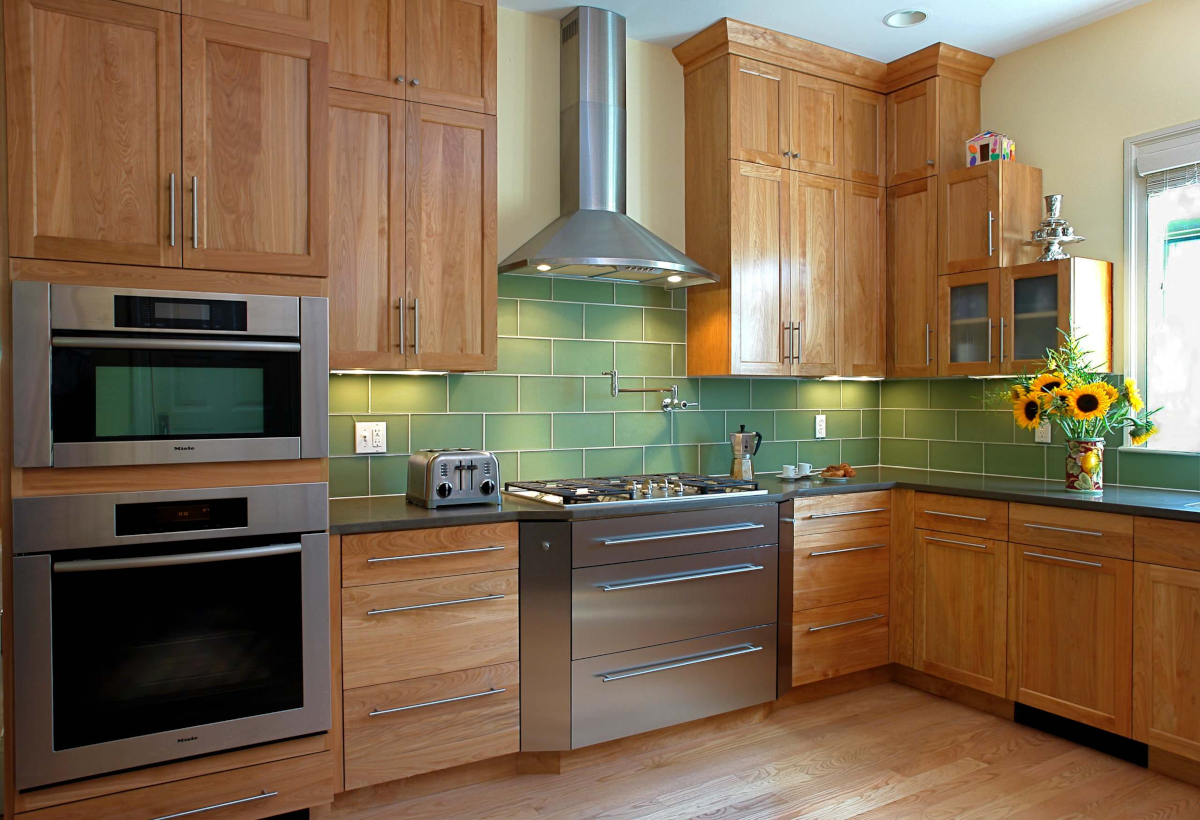 Natural wood cabinets in a kitchen with a green tile backsplash and stainless steel appliances