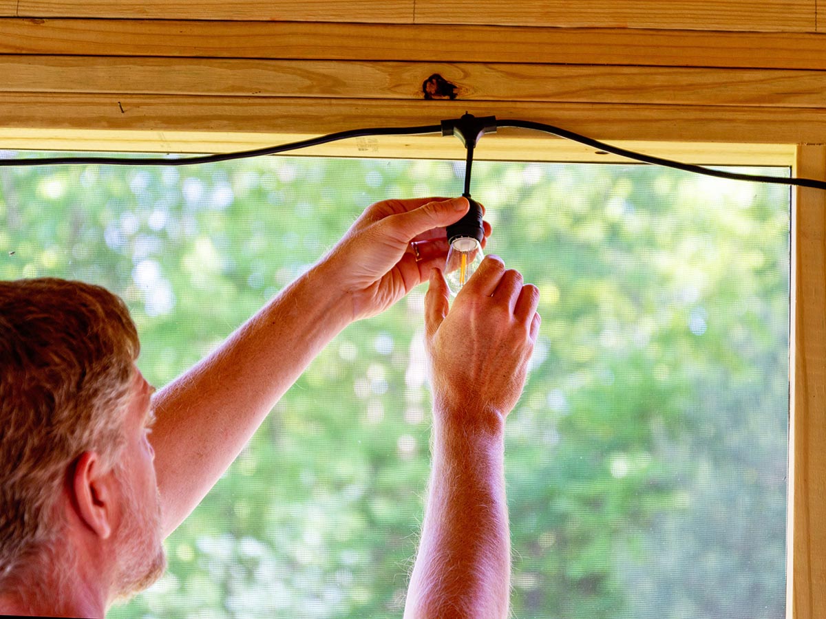 A person adjusting a light bulb on the Tiki BiteFighter LED string lights, which are hanging on a screened-in porch