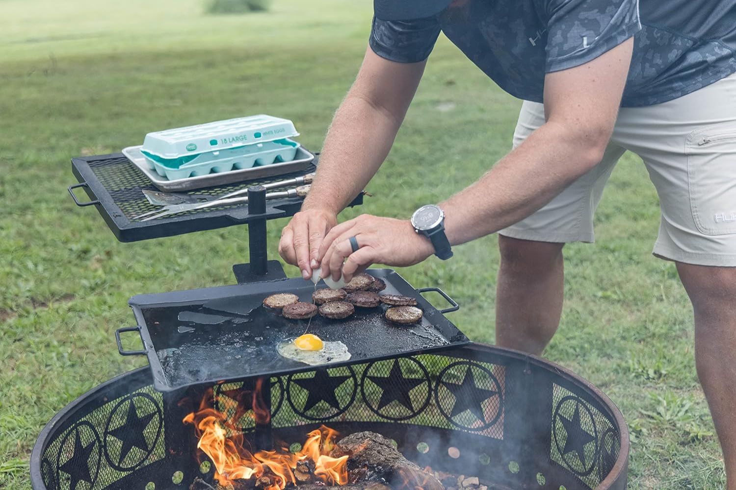 A person using the CanCooker GameMaker Gravity Grill Combo over a fire pit to cook eggs and sausage.