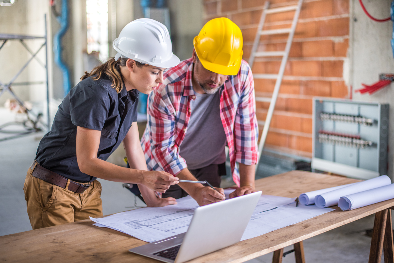 Female architect and female construction foreman looking at a blueprint while using a laptop on a construction site.