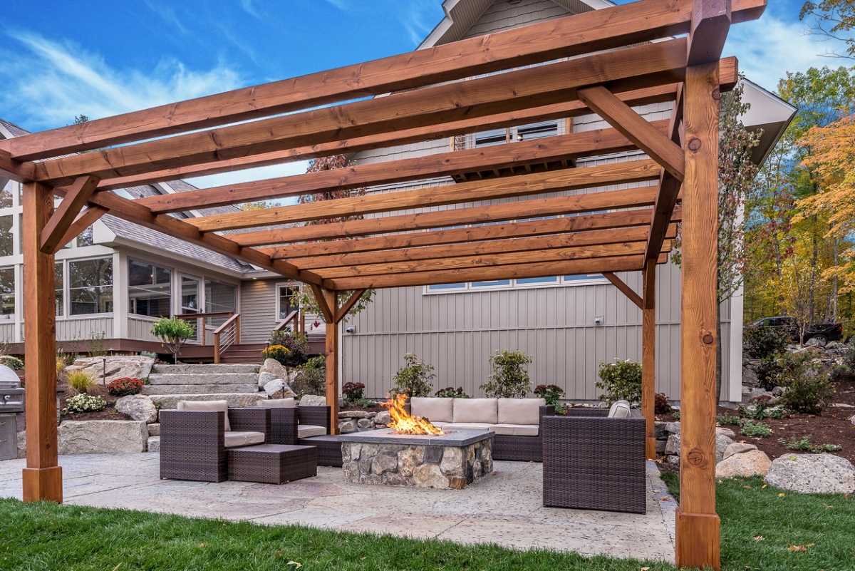 A wooden pergola is standing over a square fire pit and a matching outdoor furniture set.