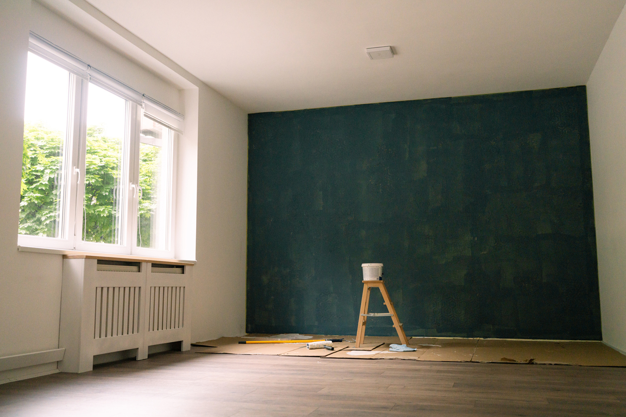 empty room with one wall painted with a thin coat of dark green paint and a ladder and paint supplies on the floor