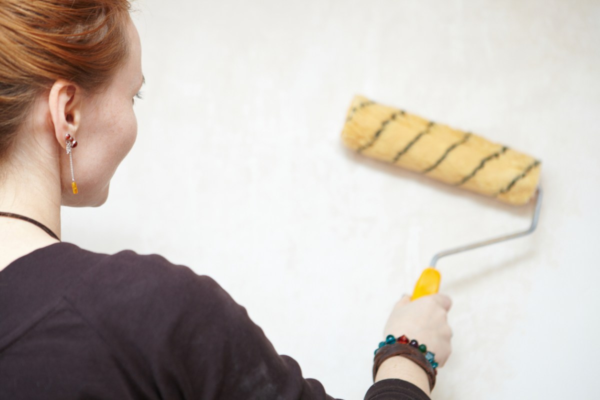 view over the shoulder of a young woman priming a white wall with a yellow paint roller