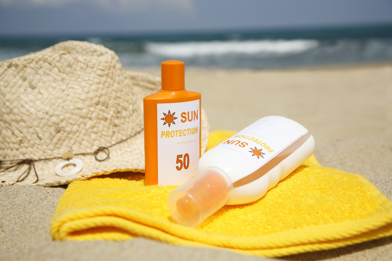 two orange and white bottles of sunscreen on top of yellow beach towel in front of straw sun hat on sand at the beach