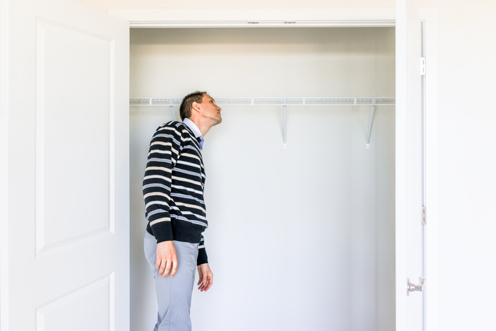 a-man-in-striped-sweater-and-grey-pants-looks-into-an-empty-bedroom-closet-against-a-white-wall