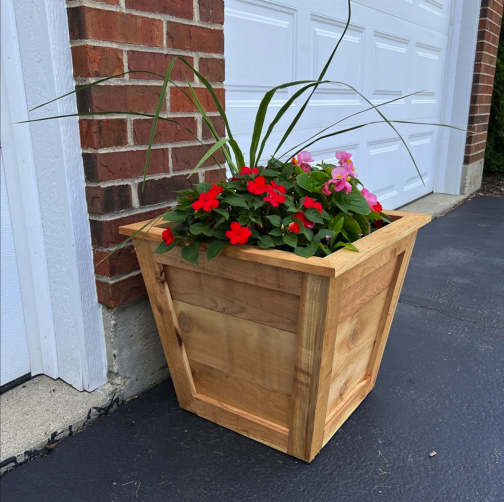 large tapered planter box made of light colored wood with red and pink flowers inside on driveway with brick garage and white garage door