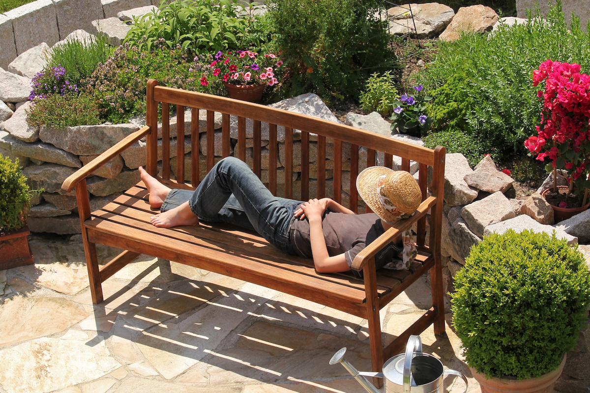 Woman laying on a wood garden bench surrounded by manicured landscape.