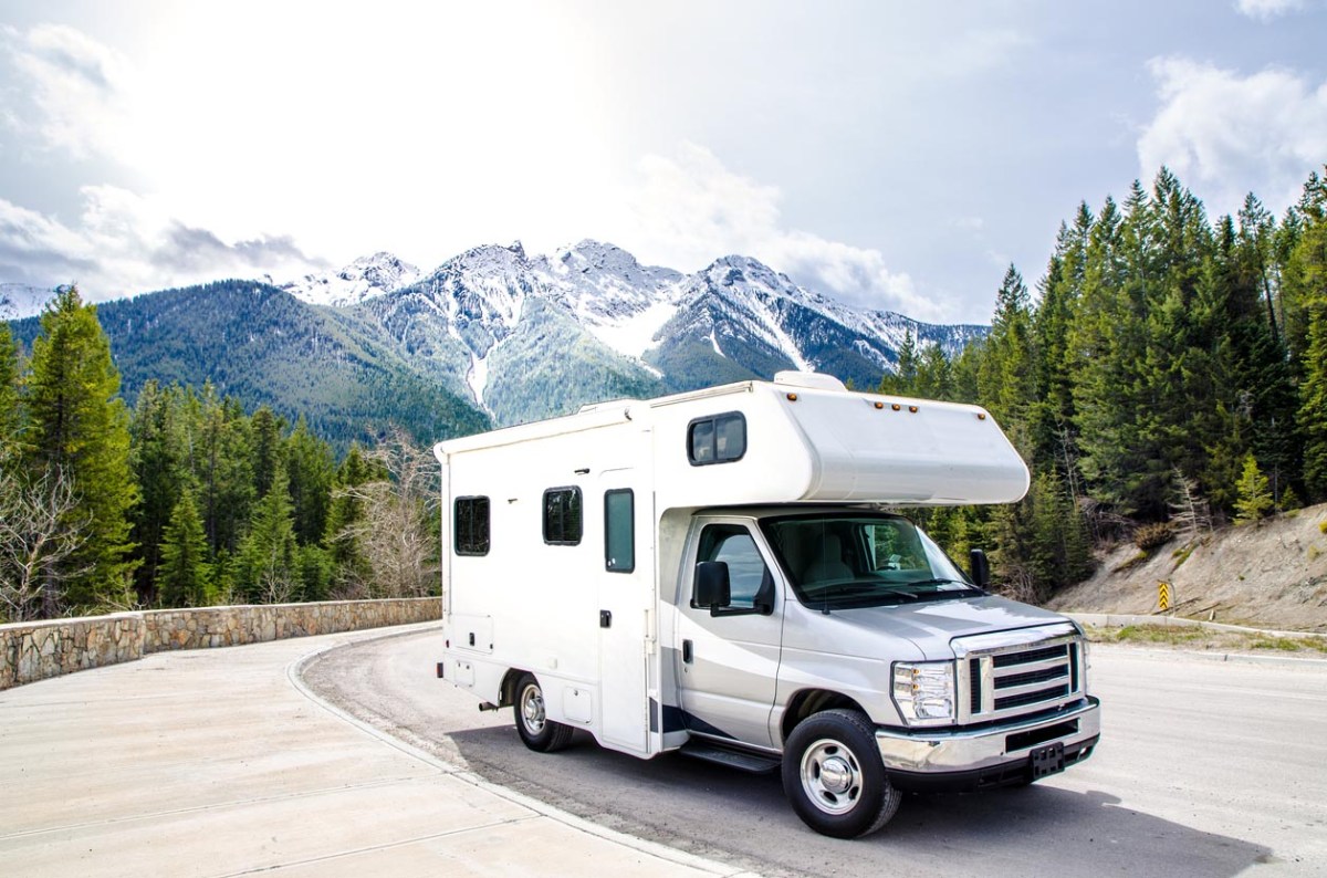 A white RV parked on a road with trees and snow-capped mountains in the background.