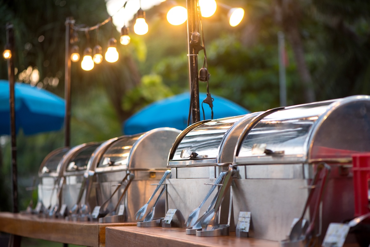 Covered food banquet trays with rolltop covers under illuminated string lights at an outdoor buffet party.