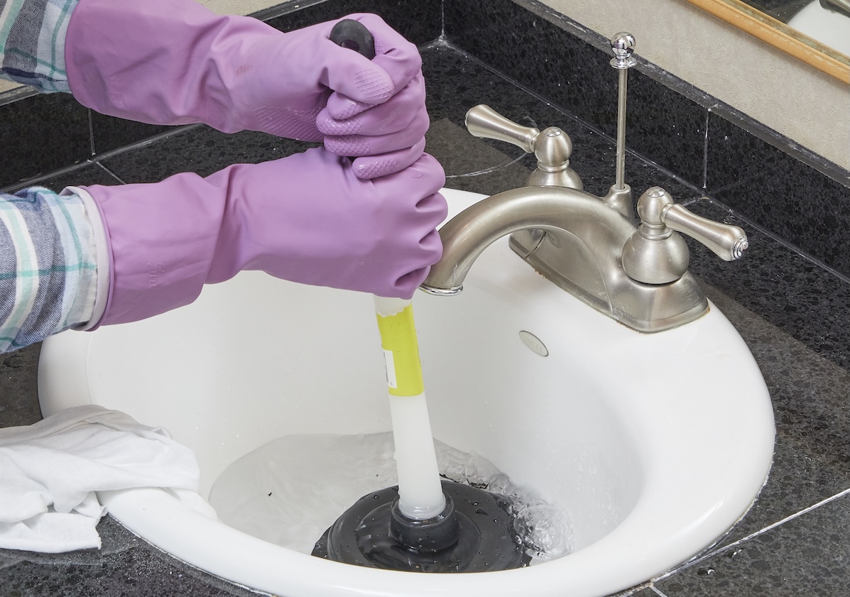 Person wearing rubber gloves plunges bathroom sink drain.