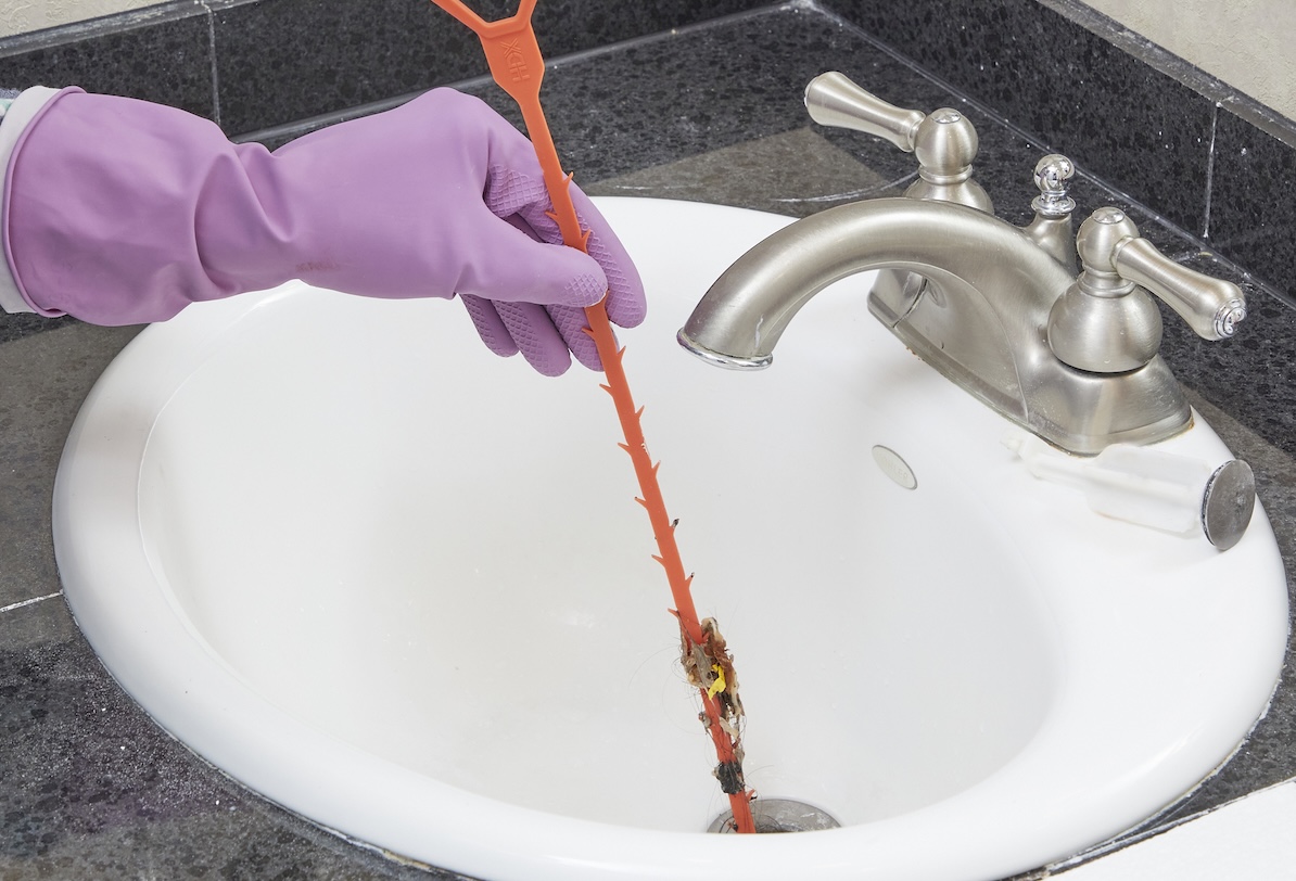 Person wearing purple rubber gloves uses a drain snake to remove debris from bathroom sink drain.