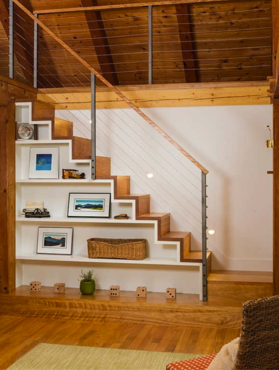 light wood staircase leading into attic space with wood beam ceilings with shelves underneath staircase displaying framed artwork