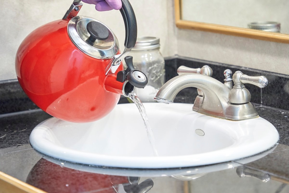 Person pours hot water from red tea kettle into bathroom sink drain.
