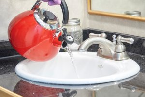 Person pours hot water from red tea kettle into bathroom sink drain.
