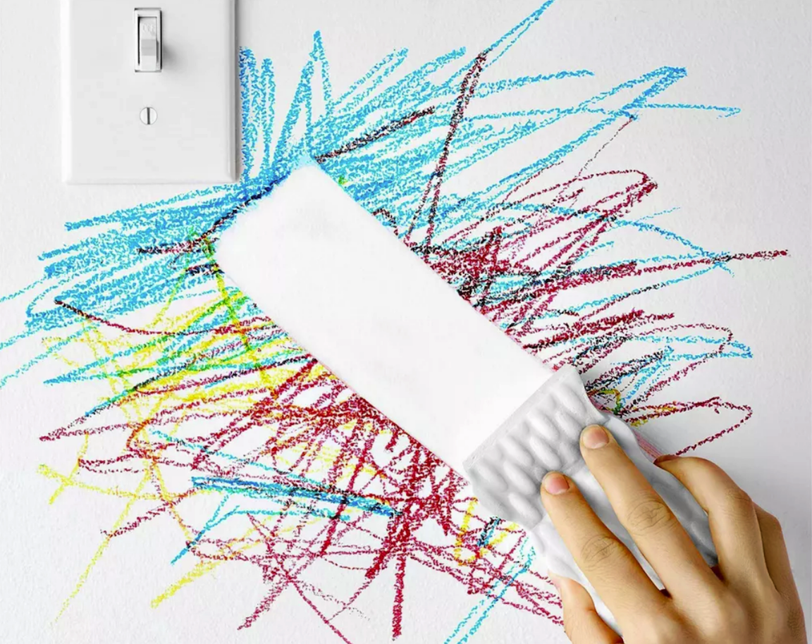 a hand drags a white magic eraser sponge across a cluster of crayon marks on a white wall next to an outlet leaving a clean white streak