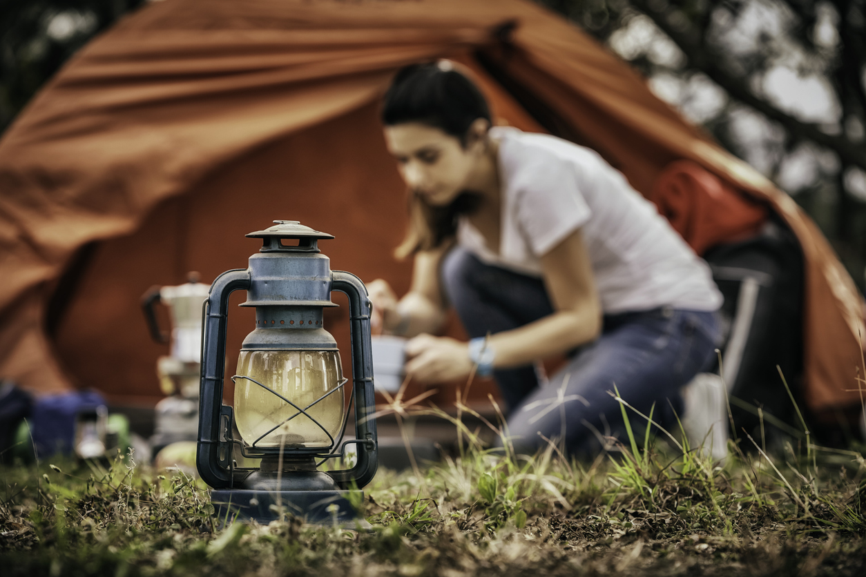 lantern in focus in front of campground where a woman has set up a red tent and is blurry in the background making a fire