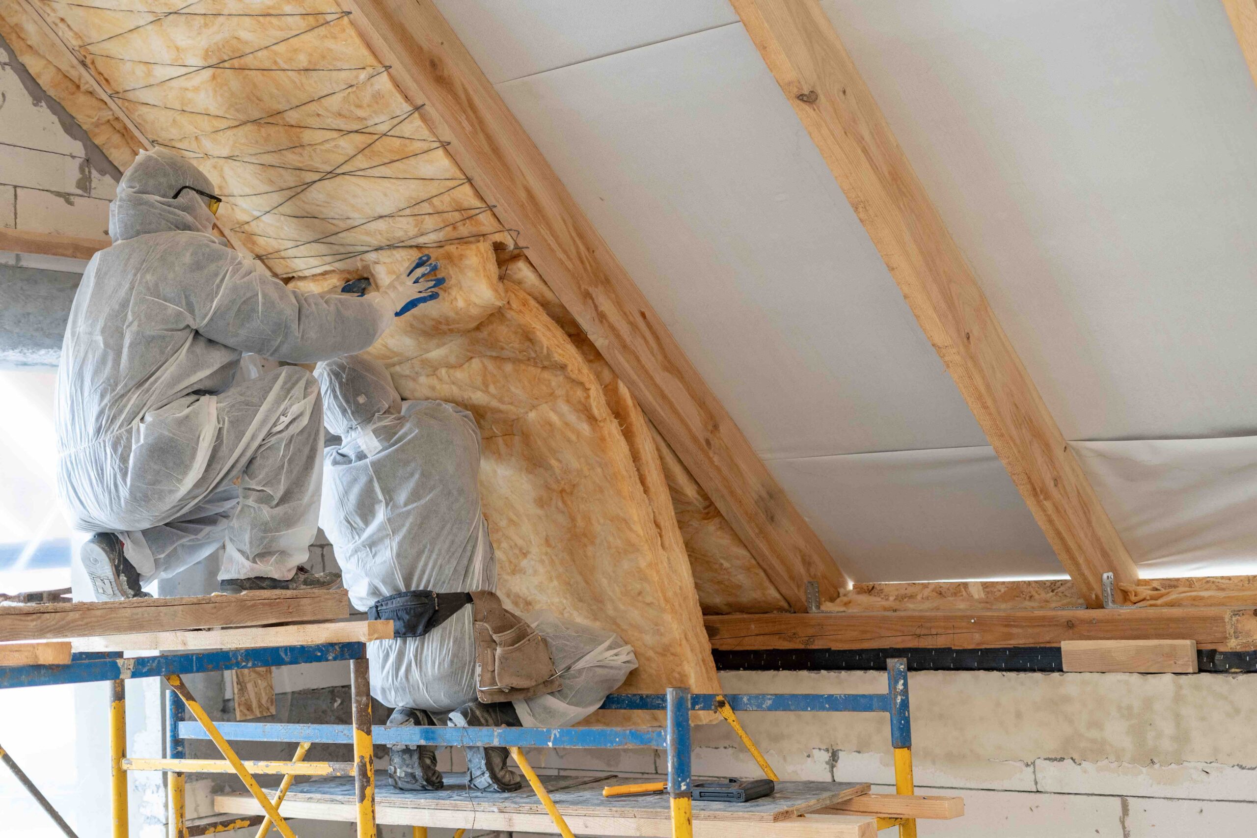 Back view of two professional worker in overalls working together with rockwool insulation, standing inside new house under construction, fasten warmth material on ceiling and wall with copy space