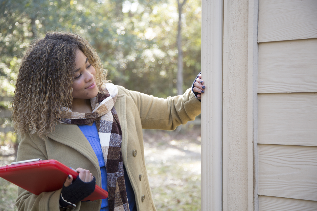 smiling woman in coat and scarf holding a red clipboard touches the beige siding on the exterior of a home to appraise it