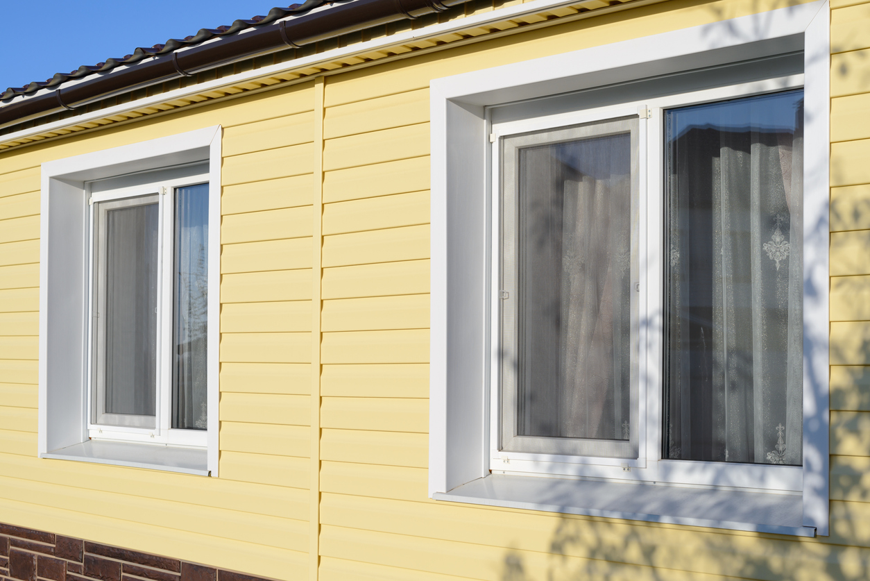 angled view of side of house with yellow vinyl siding and two windows against blue sky
