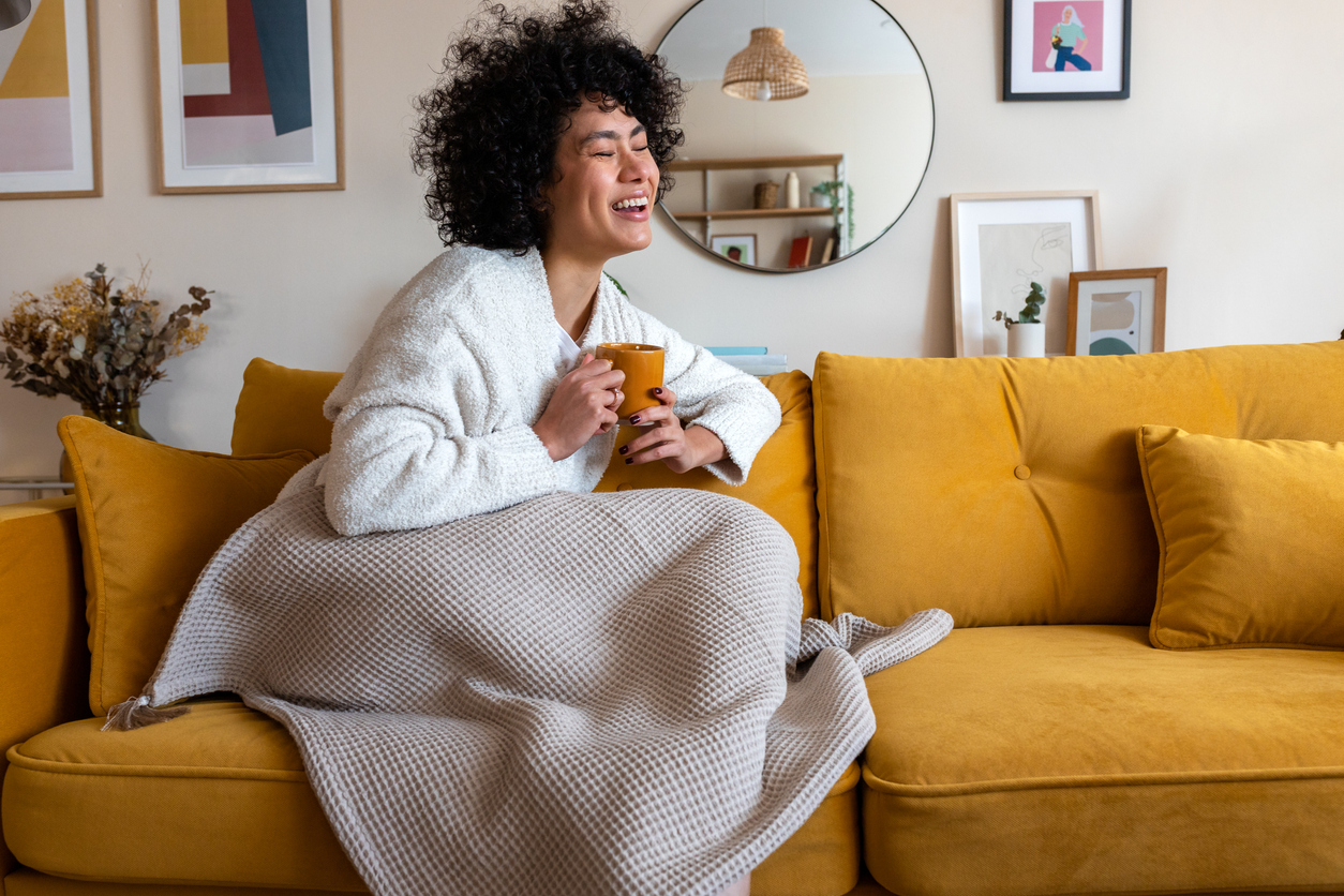 woman laughing sitting on couch covered in blanket and holding a mug