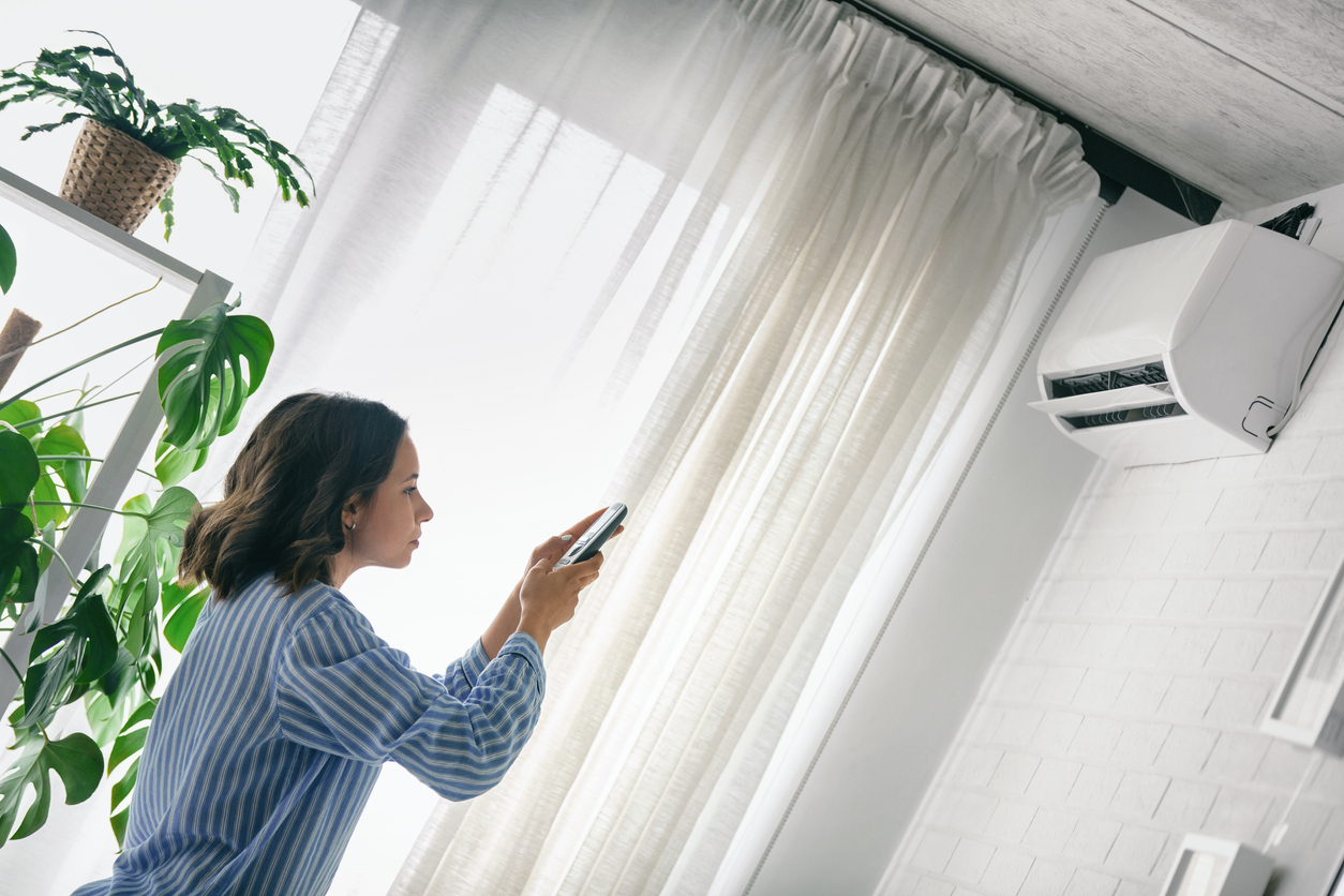young woman using remote control to operate air conditioner mounted high on the wall in light airy living room