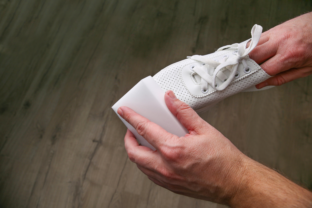 a man's hands holding a white shoe and cleaning it with a melamine sponge against the background of a dark wood floor