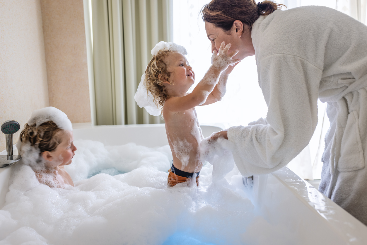 woman-in-a-white-bathrobe-leans-over-a-bubble-bath-and-plays-with-a-boy-while-another-boy-looks-on